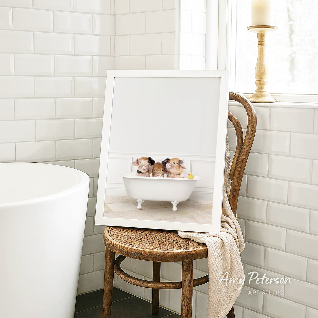 A framed image of three hamsters sitting in a bathtub, with a towel draped over the back of a wooden chair in a bathroom setting.