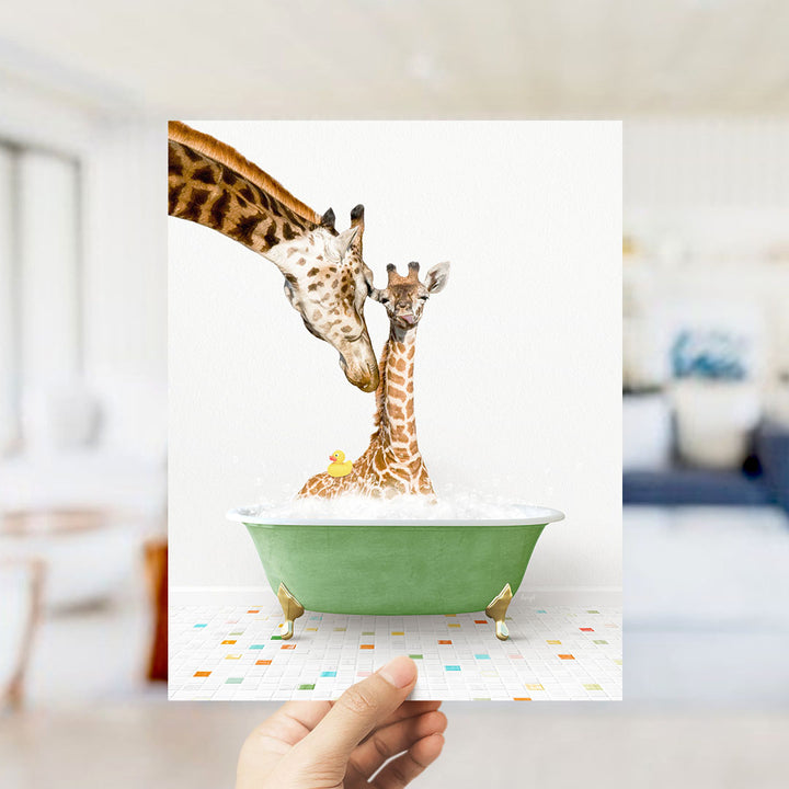 A hand holds up a framed image depicting two giraffes in a green bathtub, with one giraffe gently nuzzling the other.
