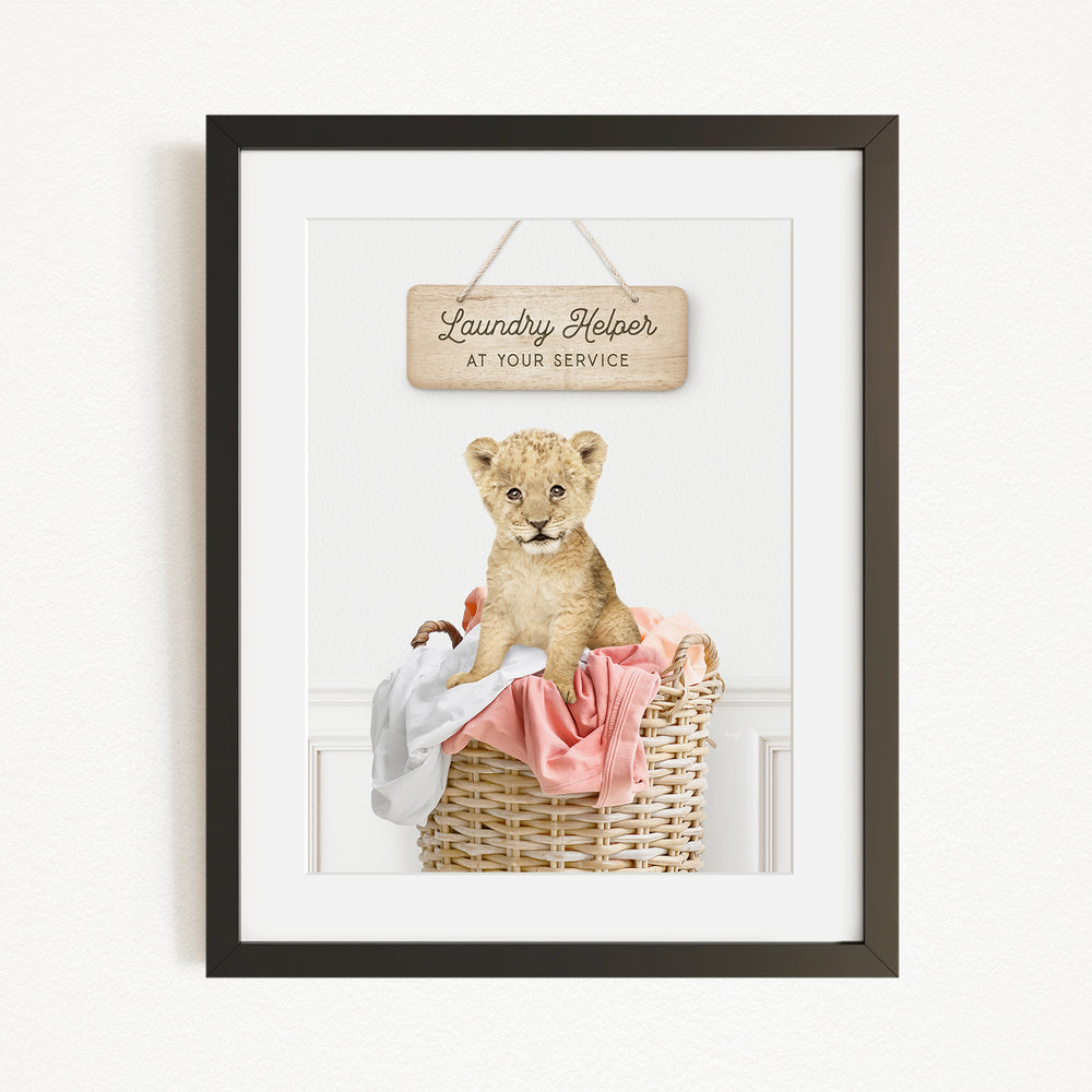 A framed image of a young lion cub sitting in a wicker basket filled with clothes, with a sign above that reads "Laundry Helper at your service".
