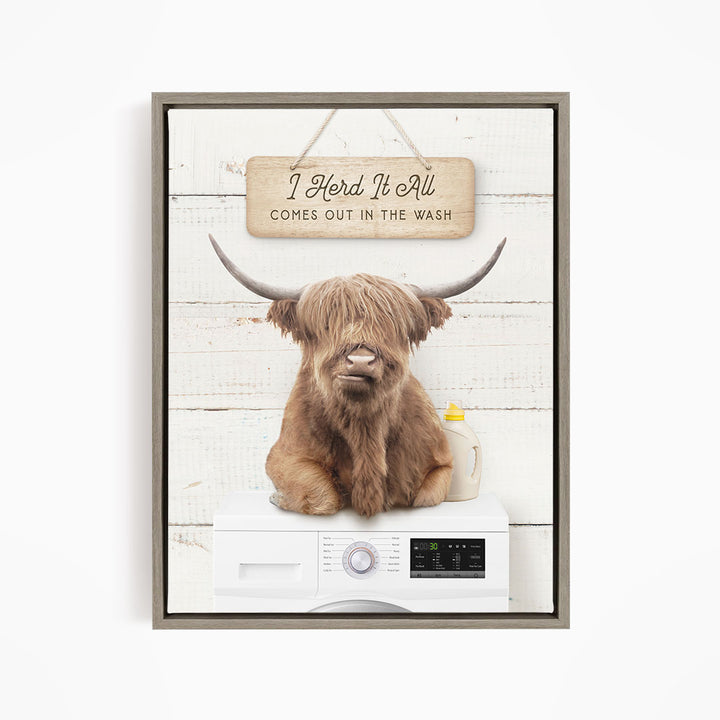 A framed image depicting a large, shaggy-haired animal, possibly a yak or a bison, sitting on top of a washing machine. The animal has horns and appears to be making a face. Above the animal, there is a wooden sign that reads "I Herd It All Comes out in the Wash".