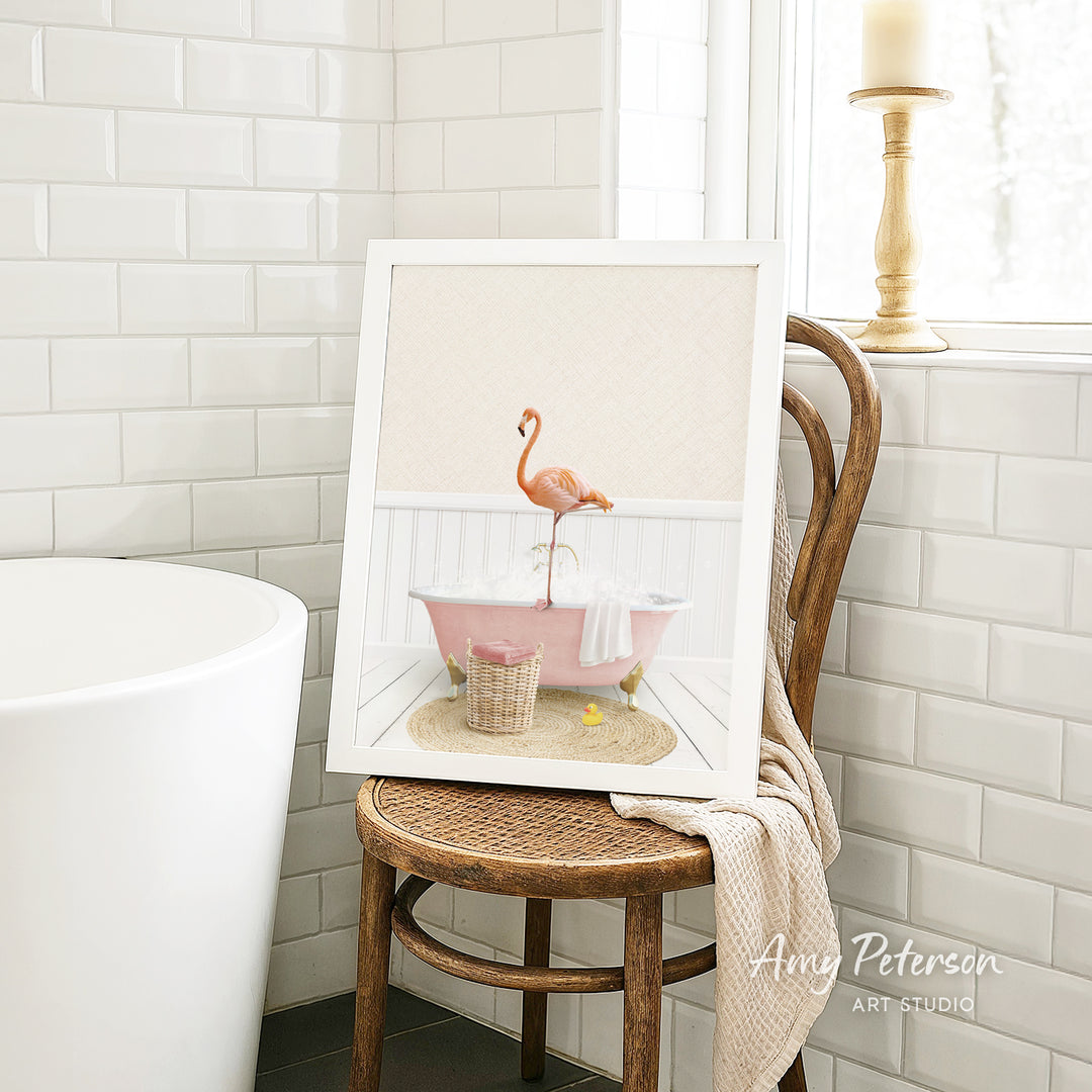 A framed image of a pink flamingo standing in a pink bathtub, surrounded by a basket of bath toys, is displayed on a wooden chair in a bathroom setting.