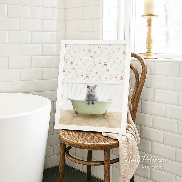 A framed image of a gray cat sitting in a green bathtub, with a floral pattern on the wall behind it, is displayed on a wooden chair in a bathroom setting.