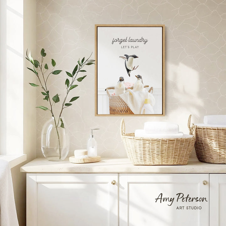 a framed poster of penguins in a basket, hanging on a wall above a white cabinet with baskets. The poster has a playful and whimsical design, and the text "Forget laundry, let's play" is displayed on it.