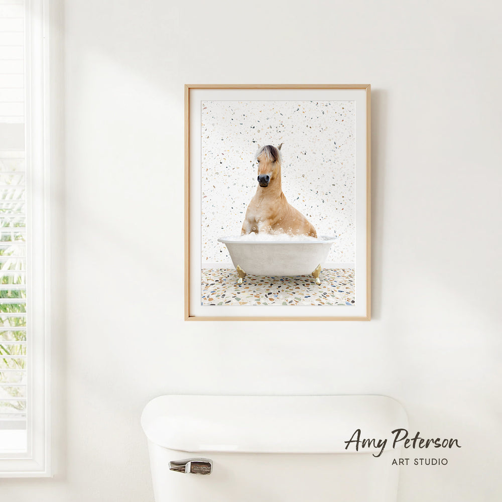 A framed image of a brown horse sitting in a bathtub, surrounded by confetti, is hanging on a white wall in a bathroom.