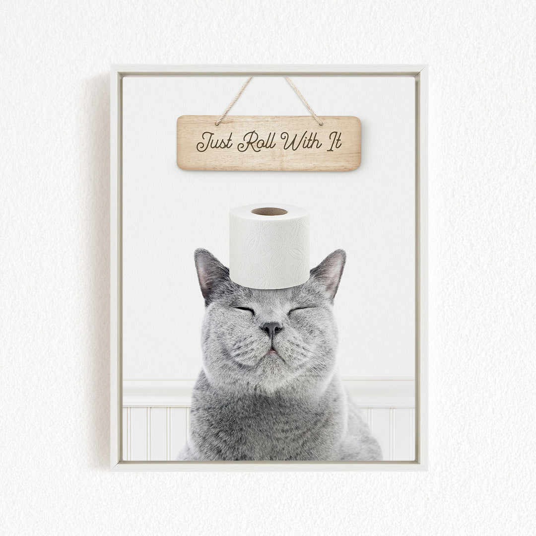 A gray cat with its eyes closed and a piece of toilet paper on its head, with a wooden sign above that says "Just Roll With It".