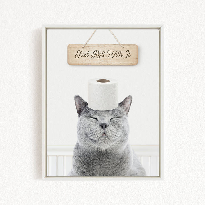 A gray cat with its eyes closed and a piece of toilet paper on its head, with a wooden sign above that says "Just Roll With It".