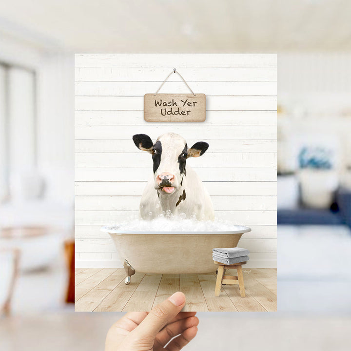 A hand holds up a framed image of a cow sitting in a bathtub, with a sign above it that says "Wash Yer Udder".