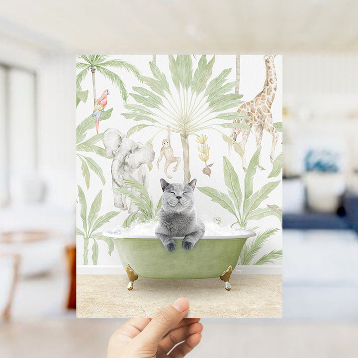 A hand holds up a framed print featuring a whimsical scene of a cat sitting in a bathtub surrounded by tropical plants and animals, including a giraffe and a parrot.