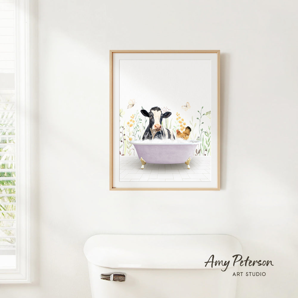 A framed print of a cow sitting in a bathtub, surrounded by flowers and butterflies, hangs on a white wall above a white toilet.