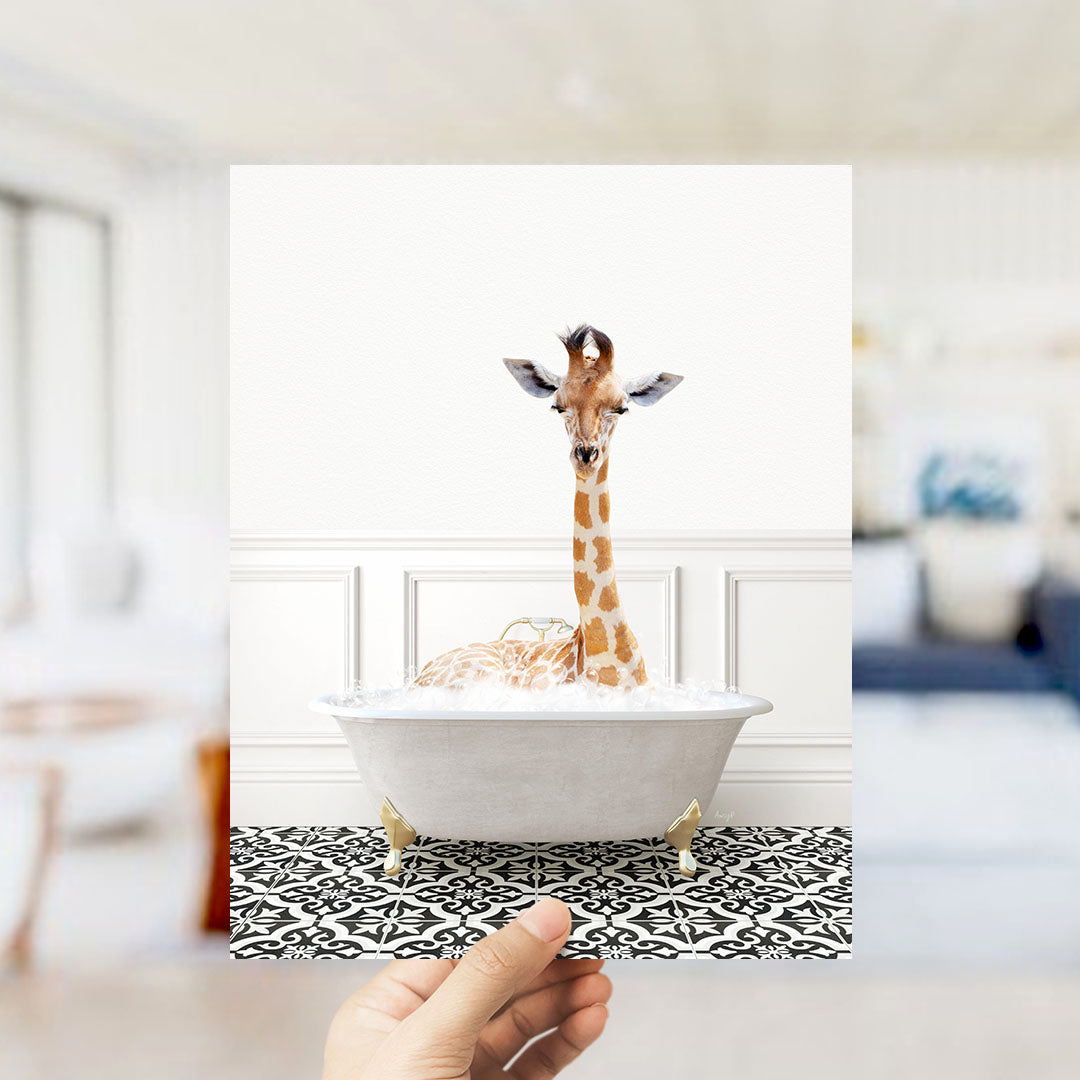 A giraffe sitting in a white bathtub filled with bubbles, with a person's hand holding the photo in the foreground.