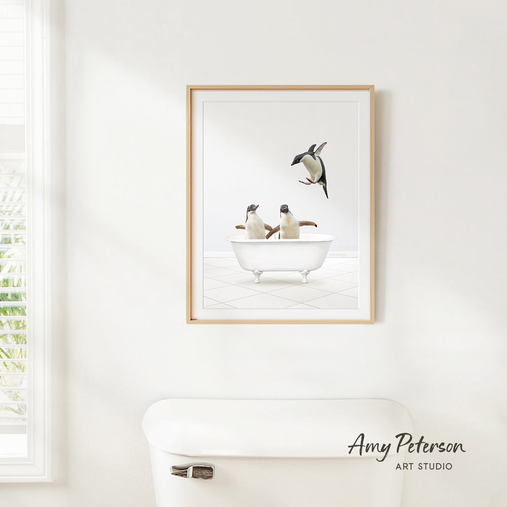 A framed image of two penguins sitting in a bathtub, with one penguin flying towards the other, is hanging on a white wall above a white toilet.
