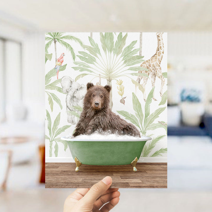 A hand holds up a framed image depicting a brown bear sitting in a green bathtub, surrounded by tropical foliage and animals.