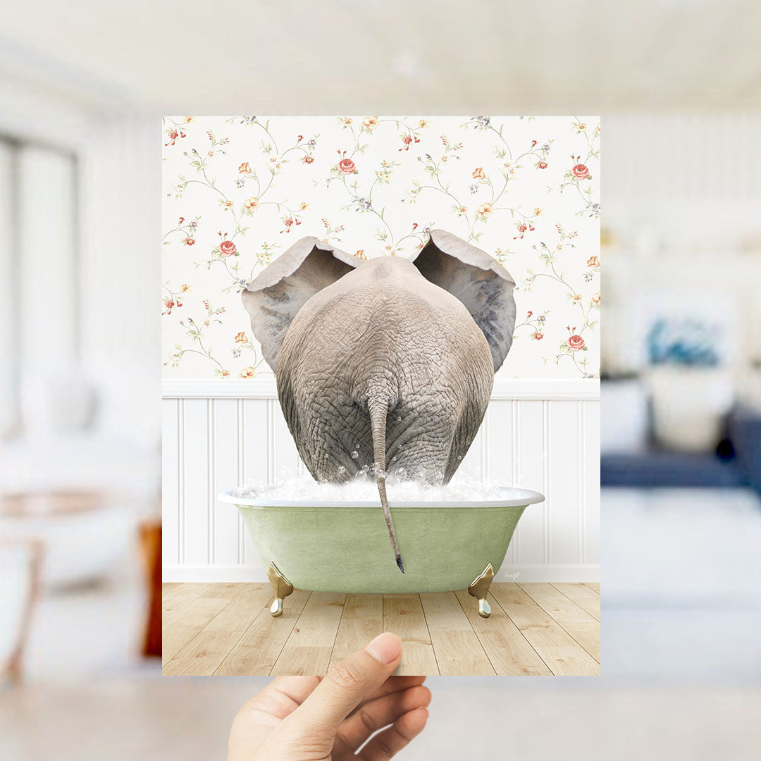 A hand holds up a framed image of an elephant standing in a green bathtub filled with water, with a floral wallpaper in the background.
