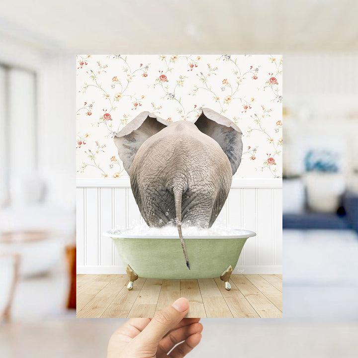 A hand holds up a framed image of an elephant standing in a green bathtub filled with water, with a floral wallpaper in the background.