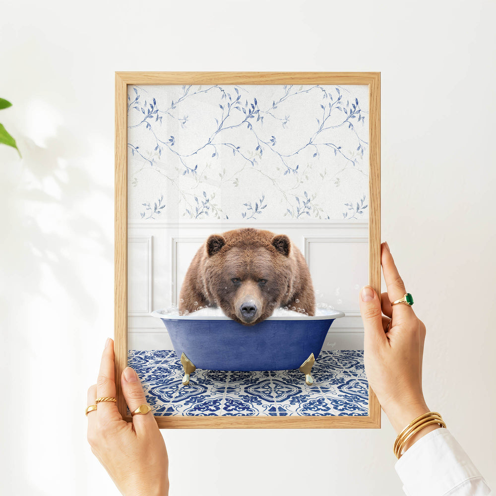 A person's hands are holding a framed image of a brown bear sitting in a blue bathtub, with a floral pattern on the wall behind the bear.