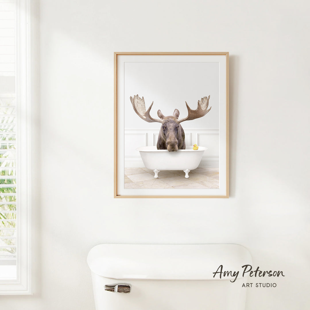 A framed image of a moose sitting in a bathtub, with its large antlers spread out, is hanging on a white wall.