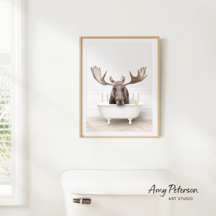 A framed image of a moose sitting in a bathtub, with its large antlers spread out, is hanging on a white wall.