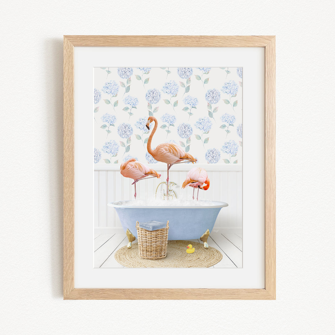 A framed image depicting three flamingos standing in a bathtub filled with water, surrounded by a blue floral pattern on the wall behind them.