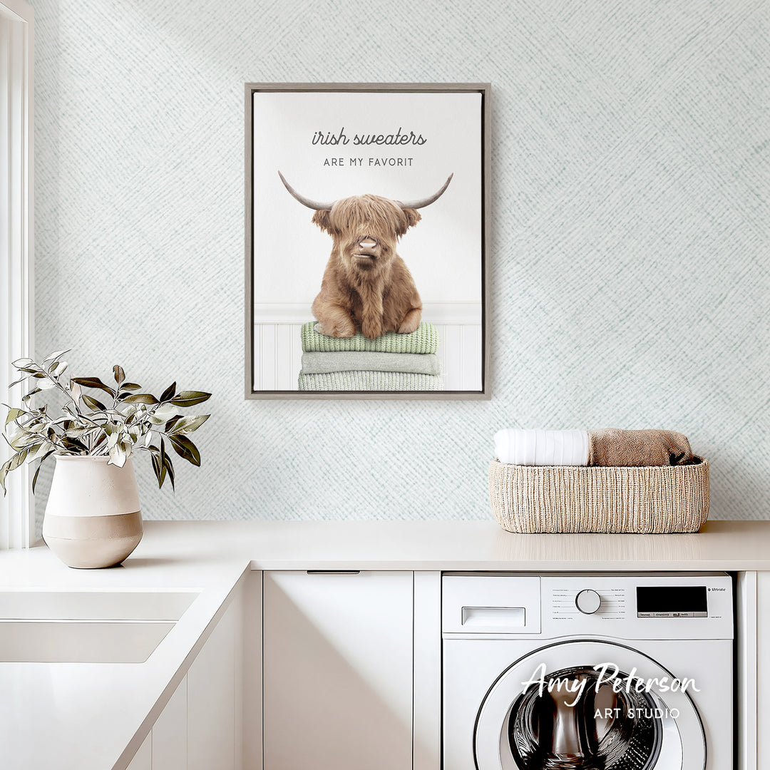 a framed poster of a Highland cow, a type of cattle, with the text "Irish sweaters are my favorite" above it. The poster is displayed on a wall above a white countertop, which has a basket of towels and a potted plant on it. There is also a washing machine visible in the background.