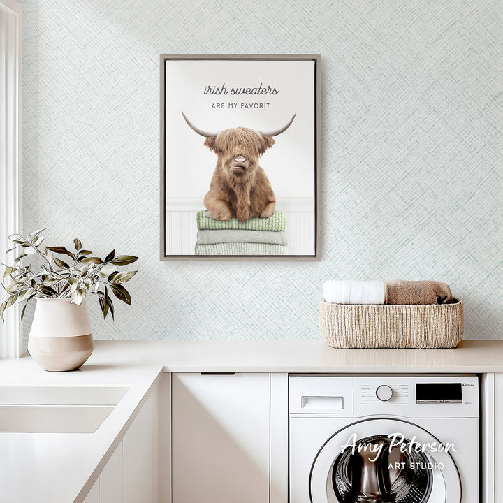 a framed poster of a Highland cow, a type of cattle, with the text "Irish sweaters are my favorite" above it. The poster is displayed on a wall above a white countertop, which has a basket of towels and a potted plant on it. There is also a washing machine visible in the background.