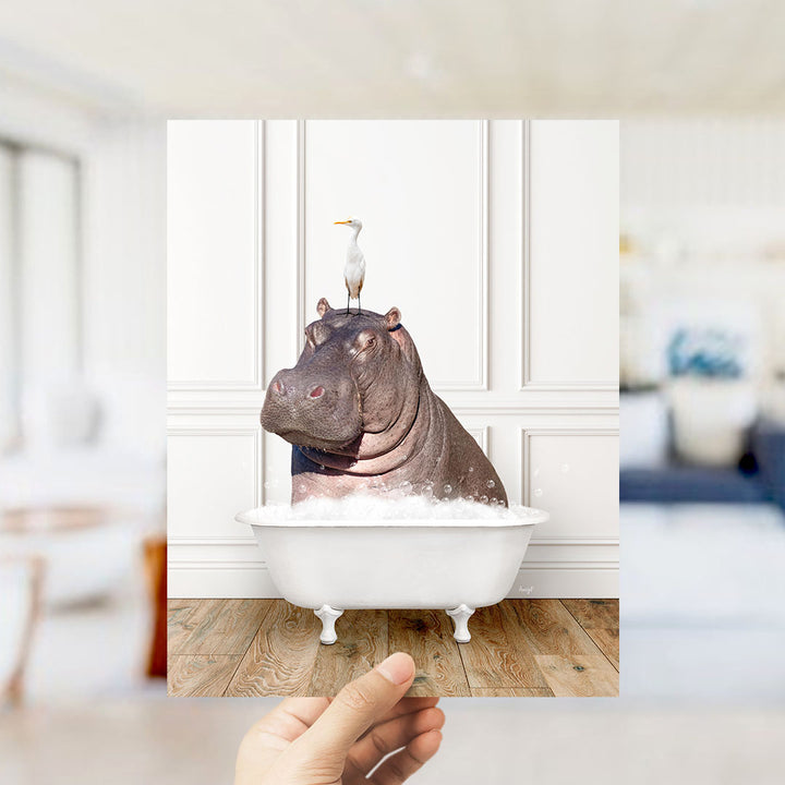 A hand holds up a framed image of a hippopotamus sitting in a bathtub, with a seagull perched on its head.