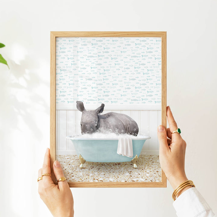 A person's hands are holding a framed picture of a rhinoceros sitting in a blue bathtub filled with bubbles, surrounded by a pattern of fish.