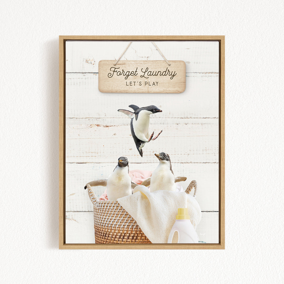 A framed image depicting three penguins in a basket, with a sign above them that says "Forget Laundry, Let's Play".