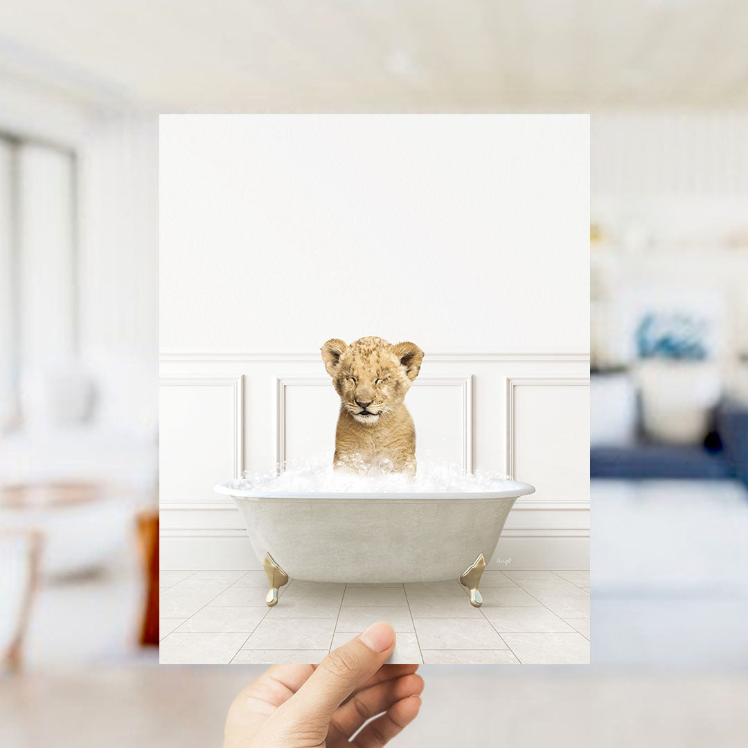 A hand holds up a framed image of a young lion cub sitting in a bathtub filled with water, with a blurred background suggesting a bathroom setting.