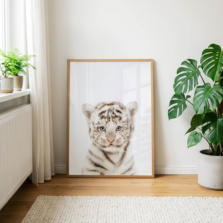 a large framed picture of a tiger cub, which is the main focus of the scene. The tiger cub is positioned in the center of the frame, and the picture is set against a white wall. The room has a wooden floor and a window with white curtains, creating a serene and natural atmosphere. There are also two potted plants in the room, one on the left side and the other on the right side, adding to the overall aesthetic.