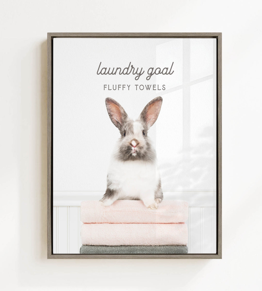 A fluffy white and gray rabbit sitting on a stack of pink towels, with the text "laundry goal FLUFFY TOWELS" displayed above the rabbit.