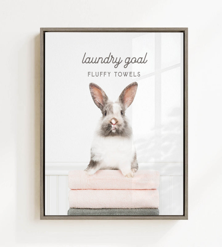 A fluffy white and gray rabbit sitting on a stack of pink towels, with the text "laundry goal FLUFFY TOWELS" displayed above the rabbit.