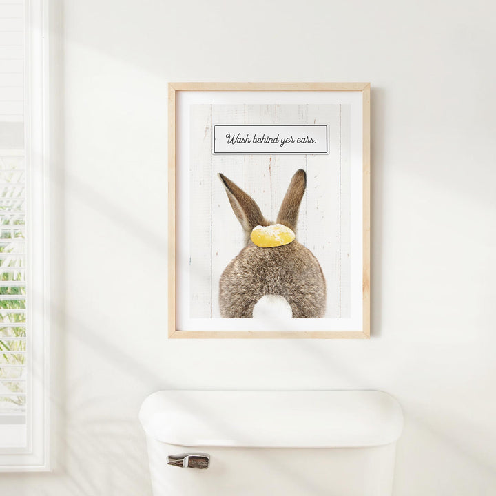 A framed picture of a rabbit with a yellow object on its head is hanging on a white wall above a white toilet.
