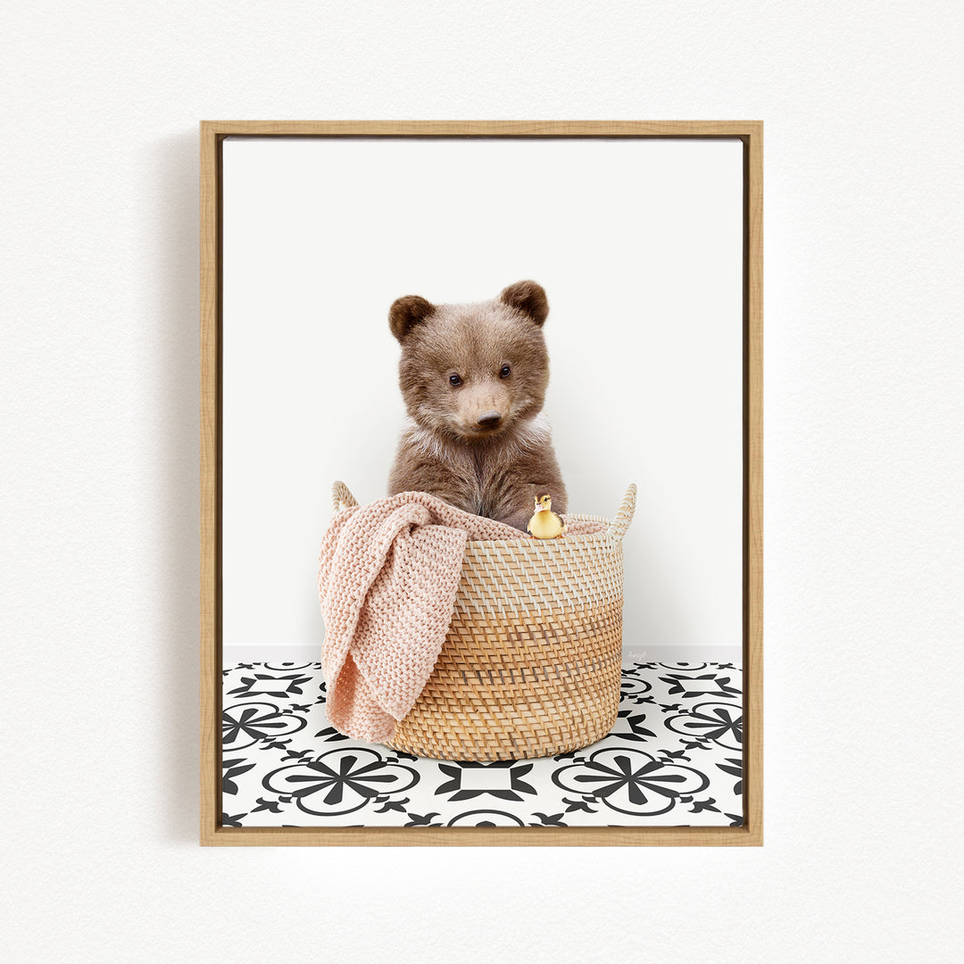 A brown bear sitting in a woven basket, with a pink blanket and a small yellow object inside the basket.