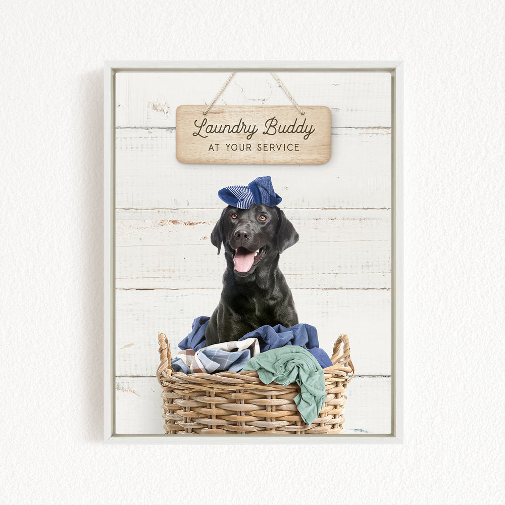 A black dog wearing a blue hat sits in a wicker basket filled with clothes, with a wooden sign above that reads "Laundry Buddy at your service".