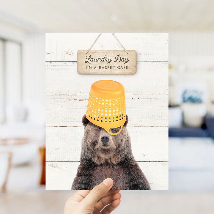 A hand holding a sign that says "Laundry Day I'm a basket case" with a bear wearing a yellow basket hat on a white background.