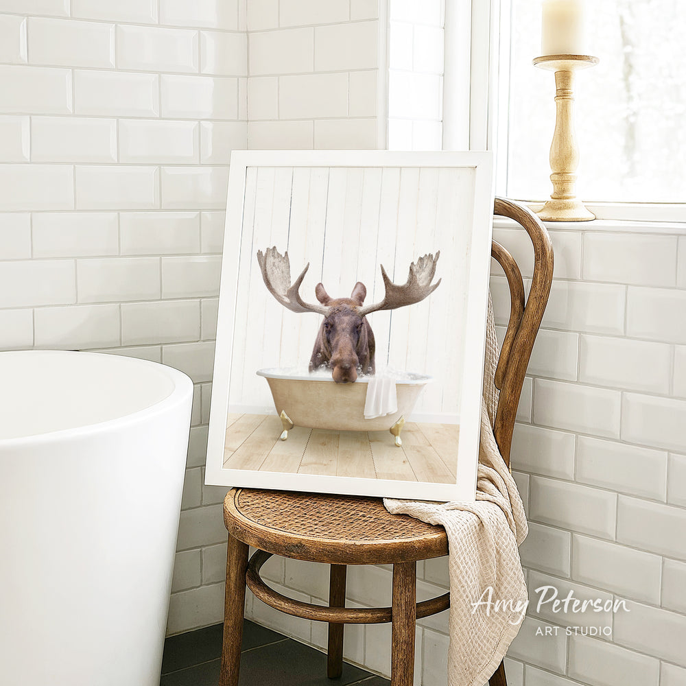 A framed image of a moose sitting in a bathtub is displayed on a wooden chair in a bathroom setting.