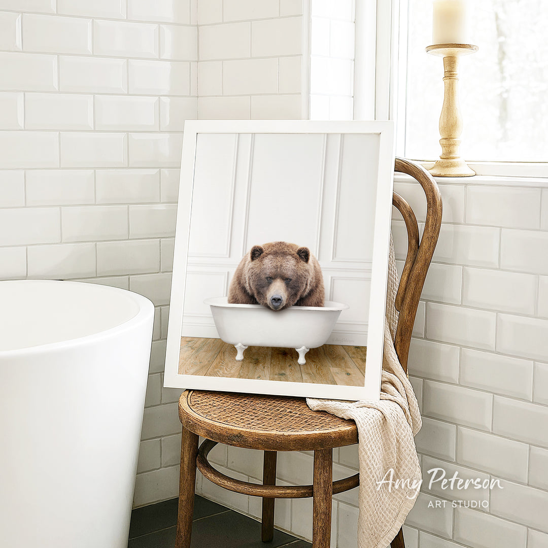 A framed image of a brown bear sitting in a white bathtub, with a towel draped over the back of the chair in a bathroom setting.
