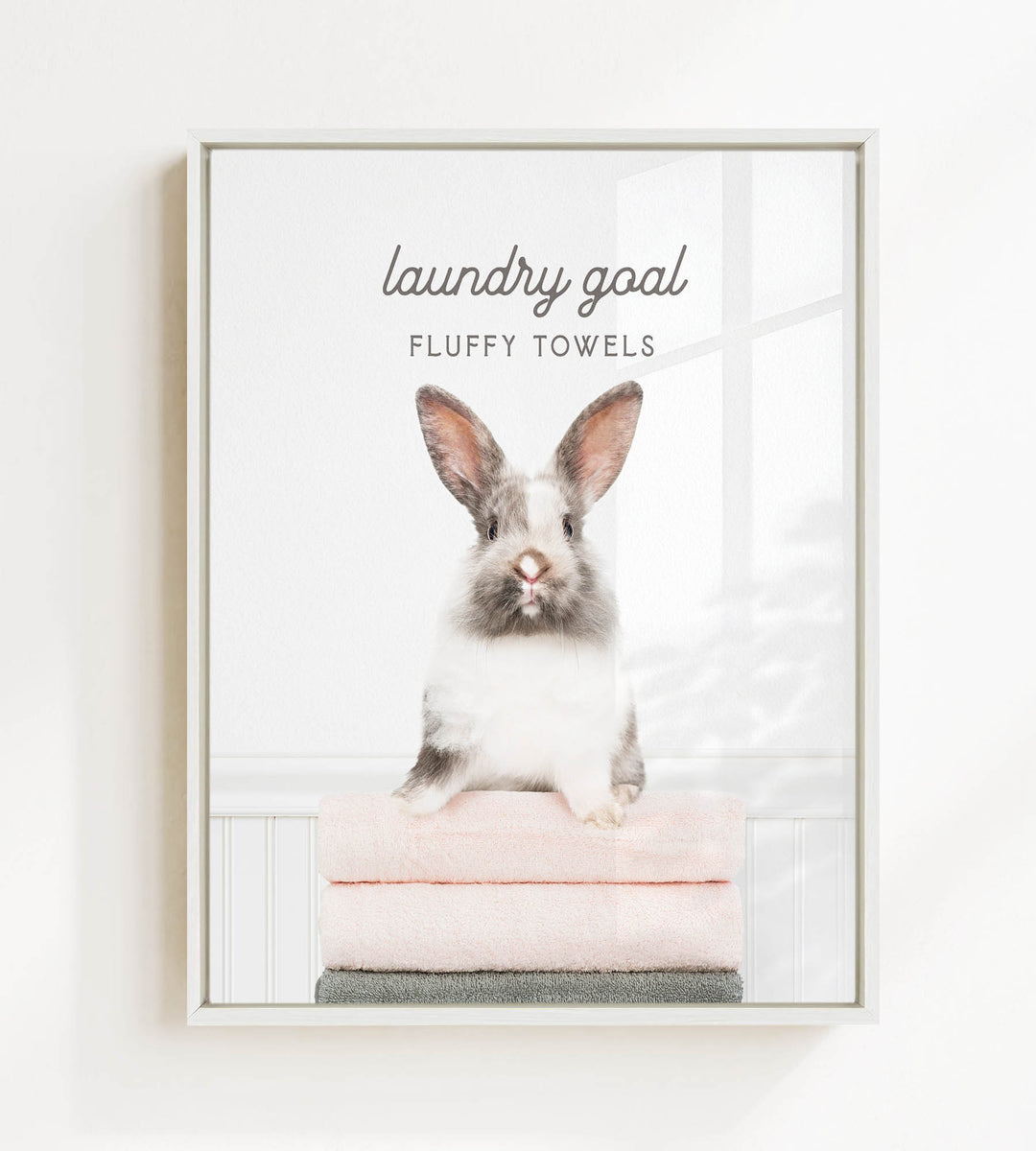 A fluffy white and gray rabbit sitting on a stack of pink towels, with the text "laundry goal FLUFFY TOWELS" displayed above the rabbit.