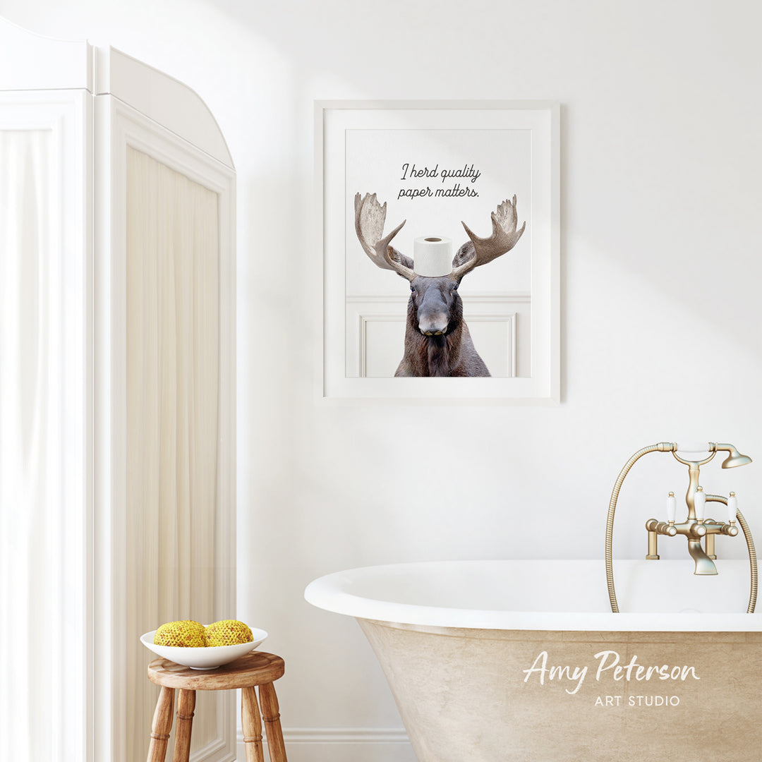 a framed picture of a moose with a roll of toilet paper on its head, hanging on a white wall above a white bathtub.
