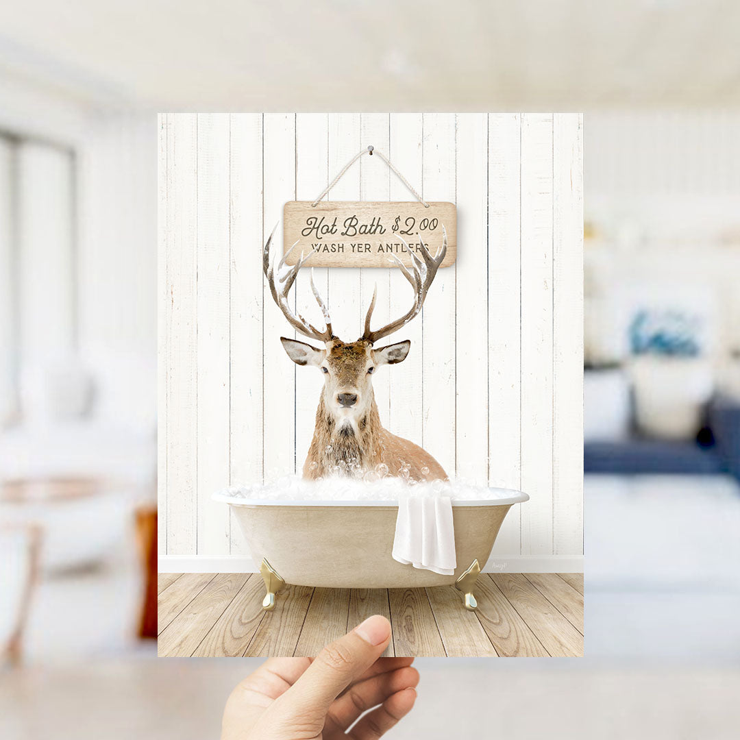 A hand holding a framed image of a deer sitting in a bathtub, with a sign above it that says "Hot Bath $2.00 Wash Your Antlers".