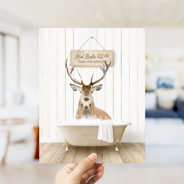 A hand holding a framed image of a deer sitting in a bathtub, with a sign above it that says "Hot Bath $2.00 Wash Your Antlers".