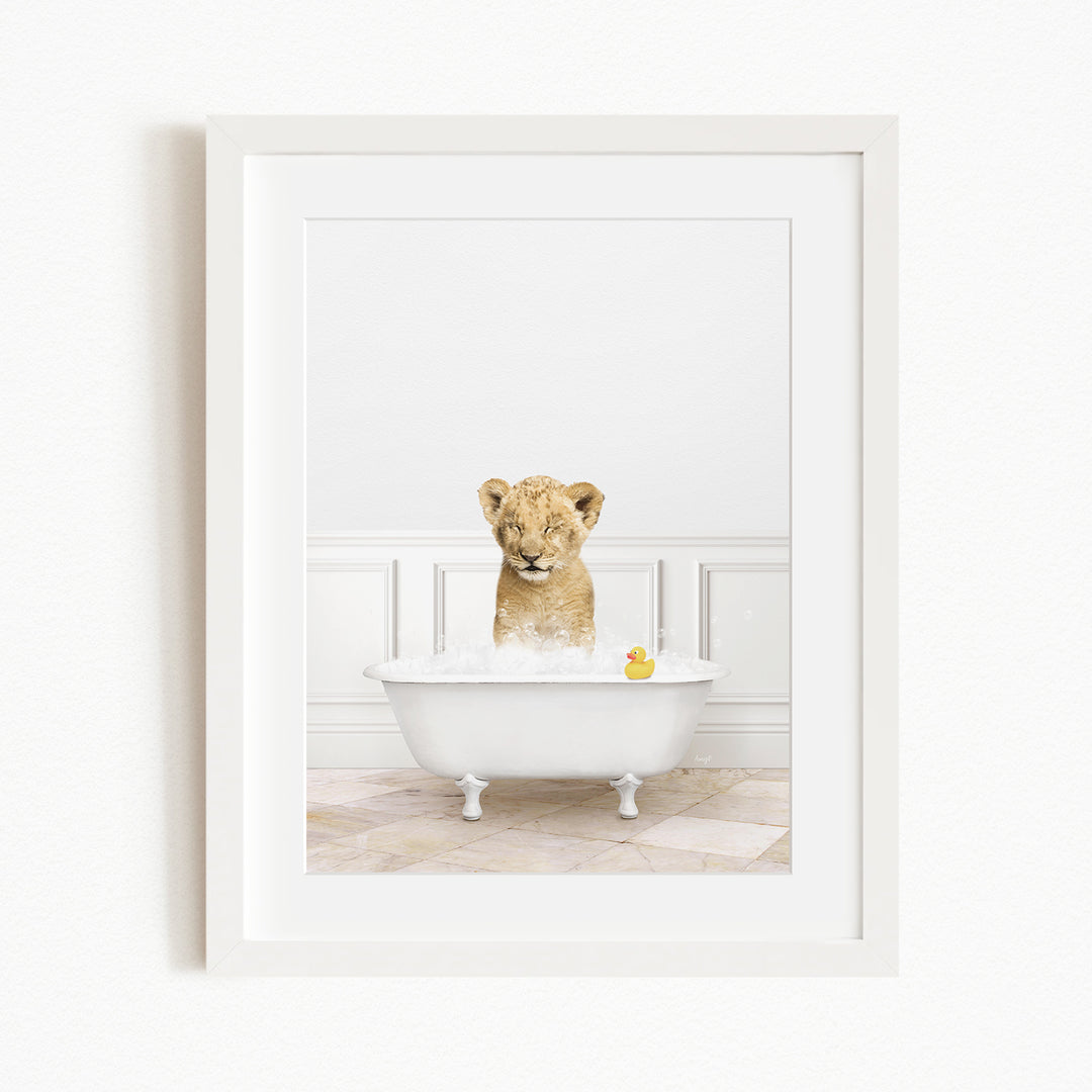 A young lion cub sitting in a white bathtub filled with bubbles, with a rubber duck floating in the water.
