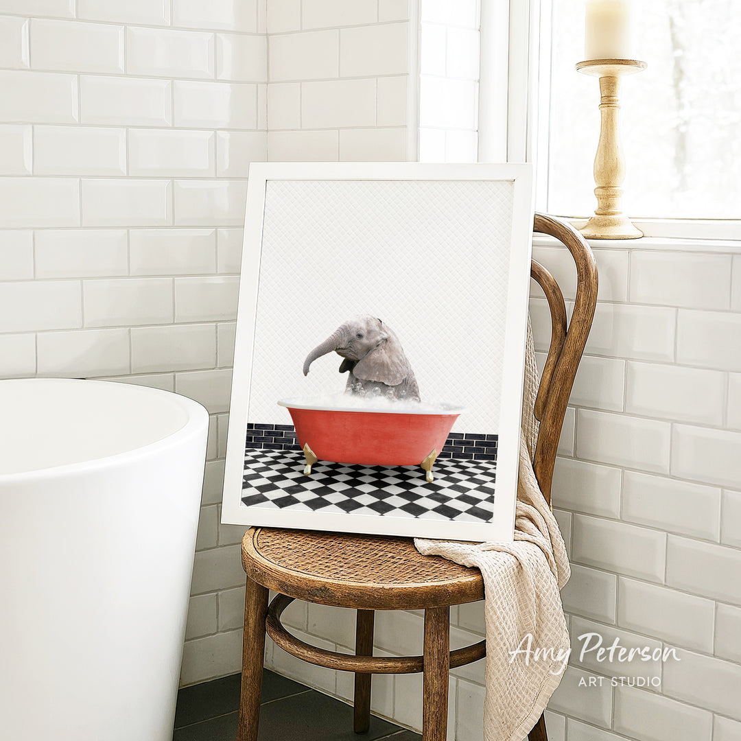 A framed image of a gray elephant sitting in a red bathtub, surrounded by a black and white checkered floor, is displayed on a wooden chair in a bathroom setting.