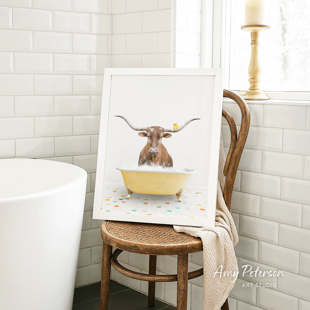 A framed image of a bull with horns sitting in a yellow bathtub, surrounded by colorful tiles, is displayed on a wooden chair in a bathroom setting.