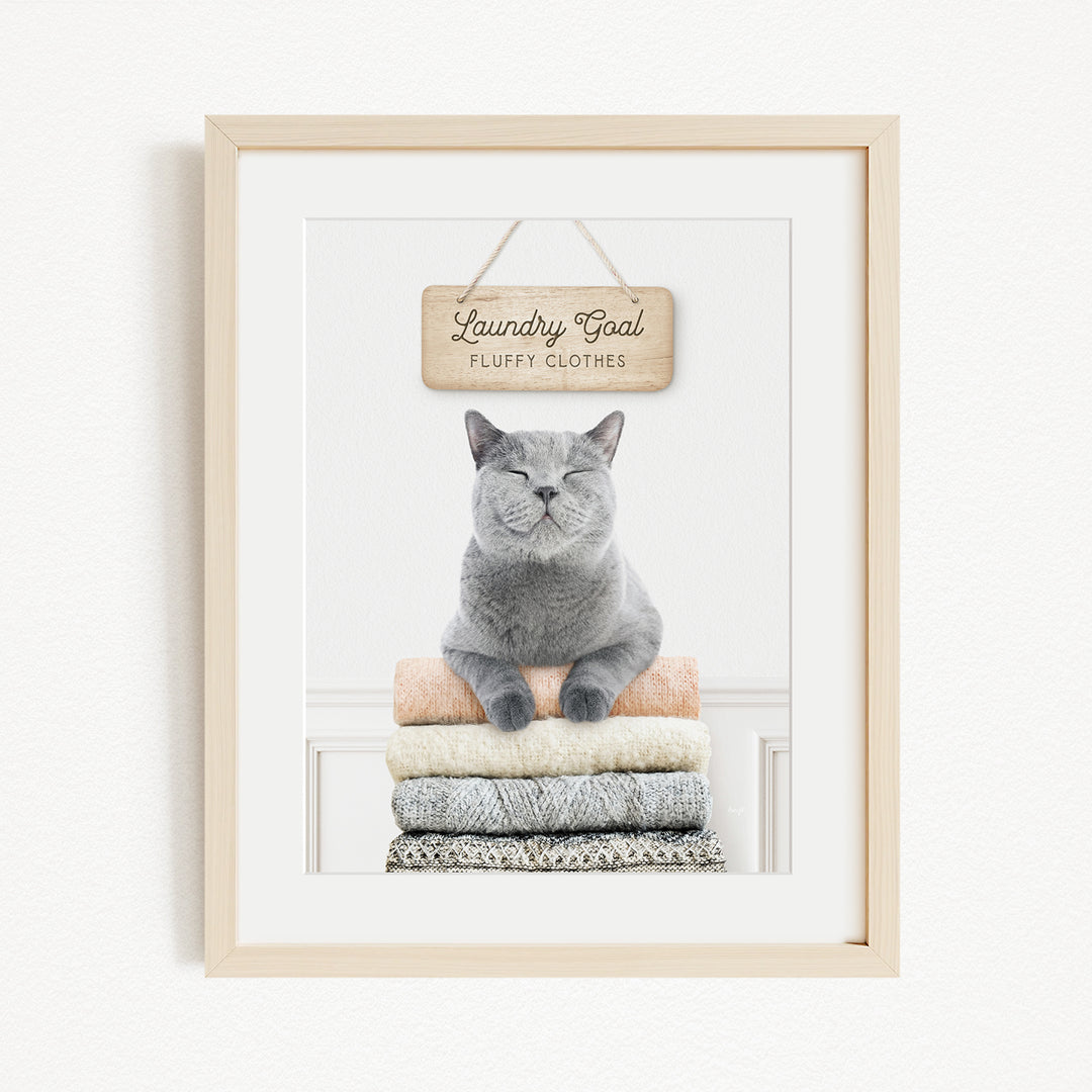 A framed image of a gray cat sitting on a stack of folded clothes, with a wooden sign above that says "Laundry Goal Fluffy Clothes".