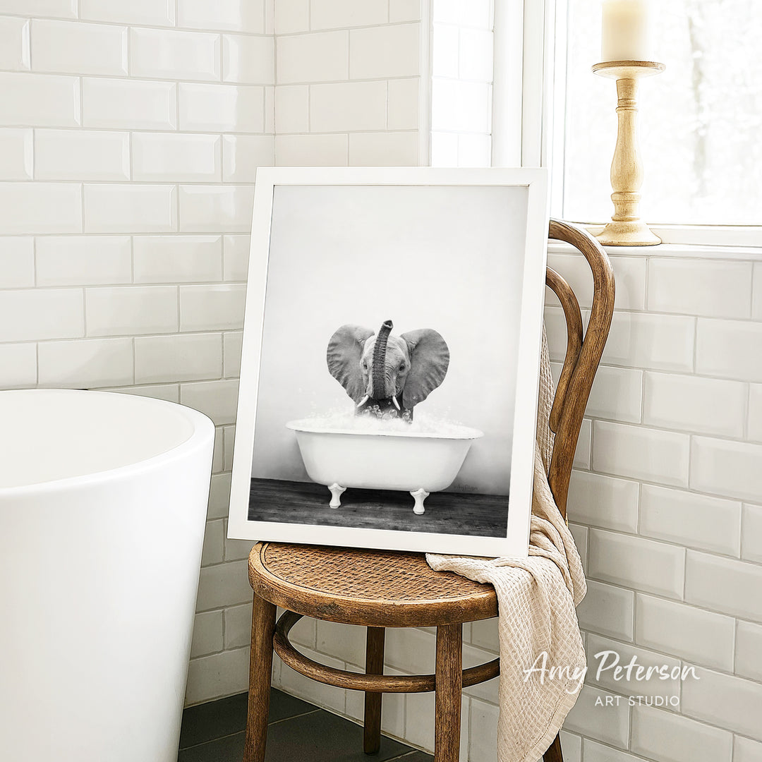 A framed black and white photograph of an elephant sitting in a bathtub, with a towel draped over the back of a wooden chair in a bathroom setting.