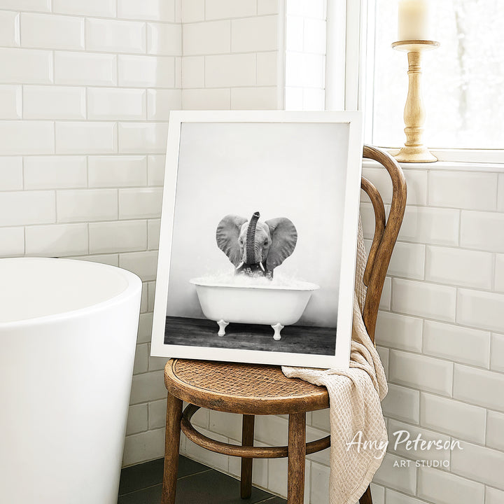 A framed black and white photograph of an elephant sitting in a bathtub, with a towel draped over the back of a wooden chair in a bathroom setting.