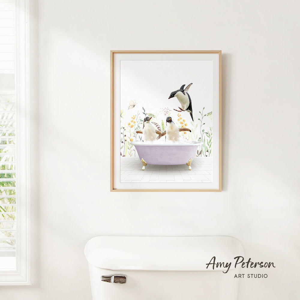 A framed illustration of two penguins sitting in a bathtub, surrounded by flowers and greenery, is hanging on a white wall.