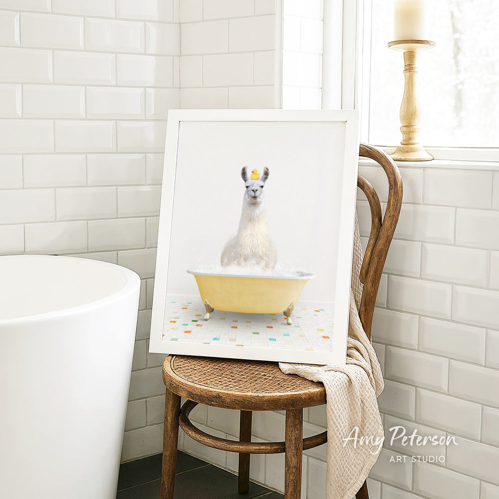 A framed image of a white llama sitting in a yellow bathtub, with a towel draped over the back of a wooden chair in a bathroom setting.