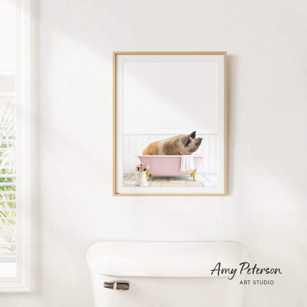 A framed image of a pig sitting in a pink bathtub, with a window and a white toilet visible in the background.